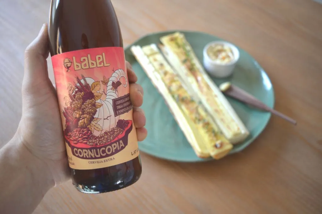 Mão segura garrafa da cerveja Cornucopia, da Babel, com palmito pupunha na brasa e manteiga ao fundo