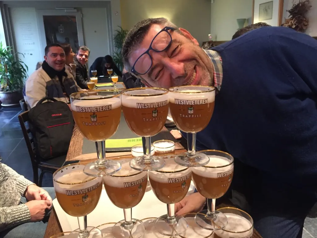 Homem de óculos sorri junto a uma pirâmide de taças Westvleteren Trappist cheias de cerveja
