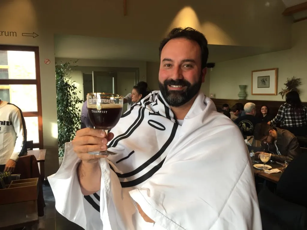 Homem sorrindo, envolto na bandeira da Expedição Cervejeira, segura taça Westvleteren com cerveja escura