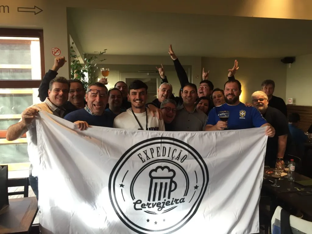 Grupo da Expedição Cervejeira posando com a bandeira do grupo e copos de cerveja em um café na Bélgica