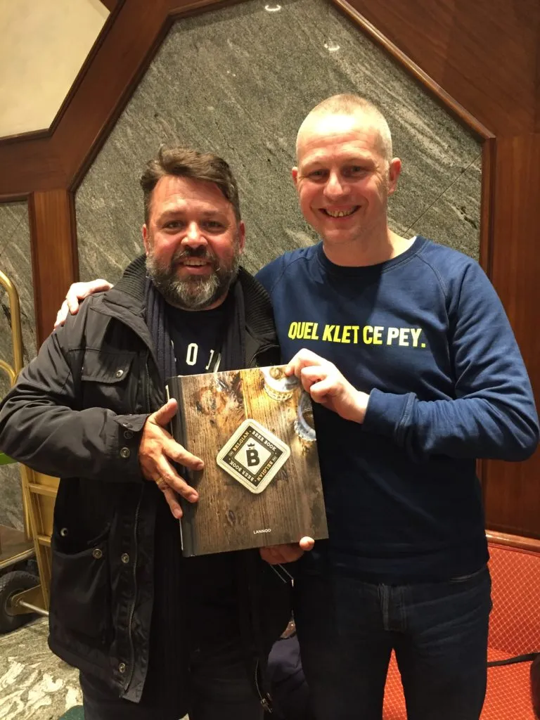 Dois homens sorriem segurando o livro Belgian Beer Book, em saguão com parede de pedra