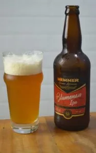 Garrafa e copo de cerveja Hemmer Blumenau IPA, de cor dourada e espuma branca, sobre mesa de madeira