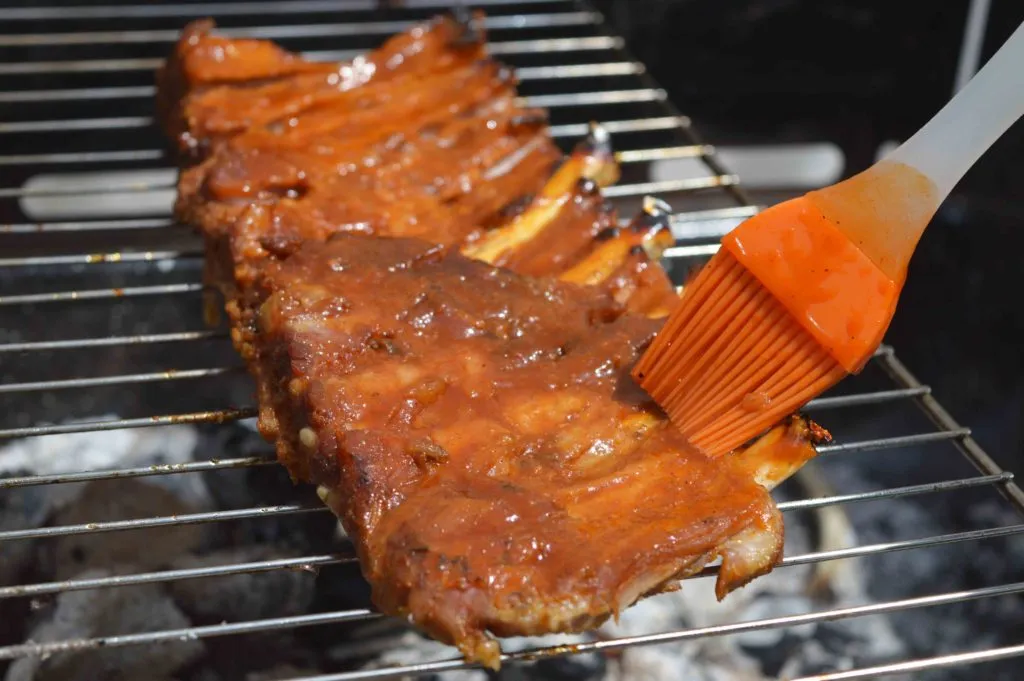 Costelinha na grelha sobre brasas sendo pincelada com molho barbecue por pincel de silicone laranja