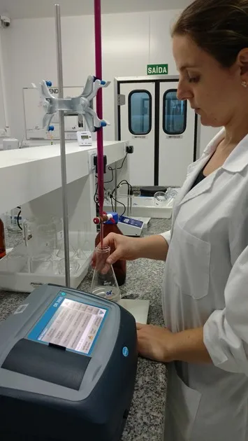 Mulher de jaleco analisa uma amostra de água em laboratório, com um equipamento de bancada e bureta ao lado