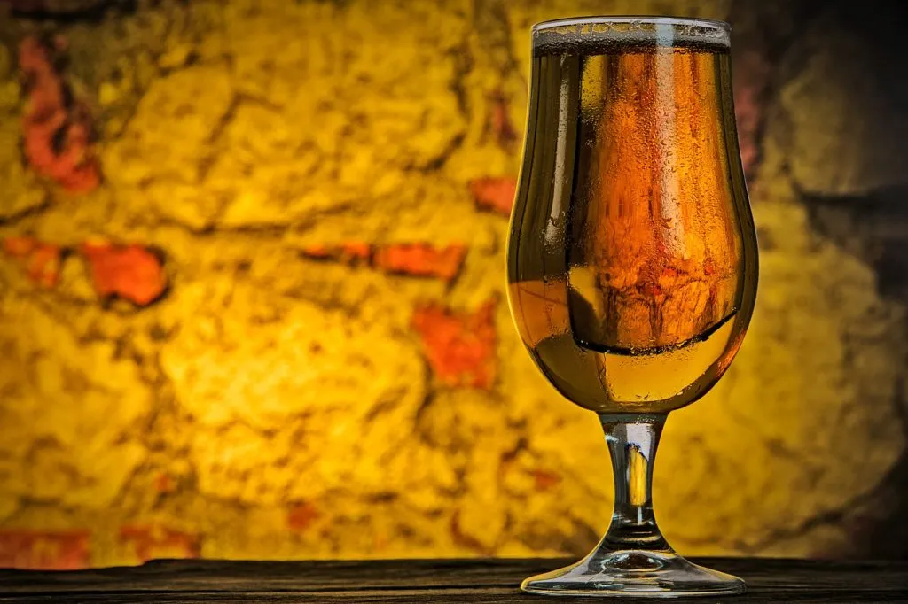 Taça de cerveja dourada com colarinho fino sobre bancada de madeira, diante de parede de pedra amarelada