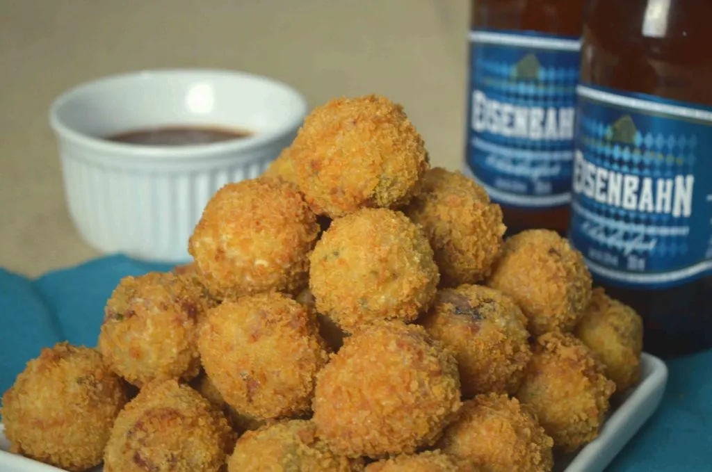 Pilha de bolinhos de aipim empanados e dourados, com molho em ramequim e garrafas Eisenbahn ao fundo