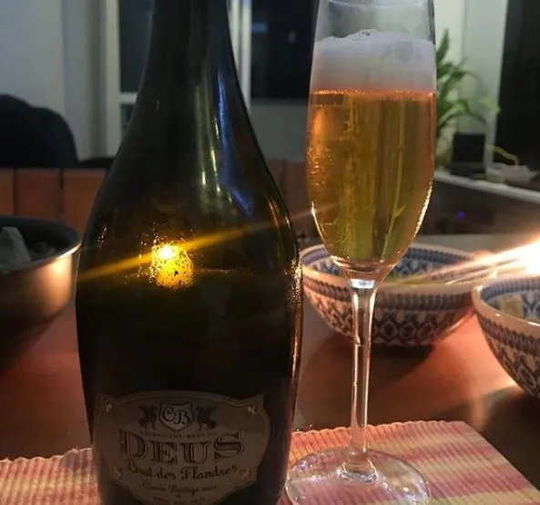 Garrafa Deus Brut des Flandres ao lado de taça flûte com cerveja dourada e espuma, sobre a mesa do jantar