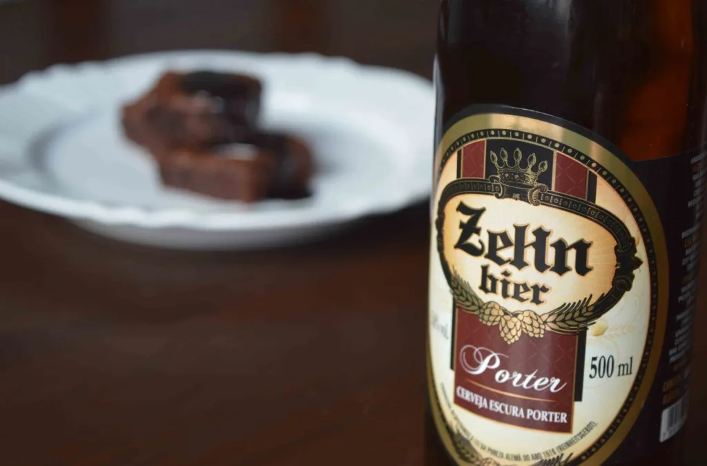 Garrafa de cerveja Zehn Bier Porter em primeiro plano, com brownies em prato branco desfocado ao fundo