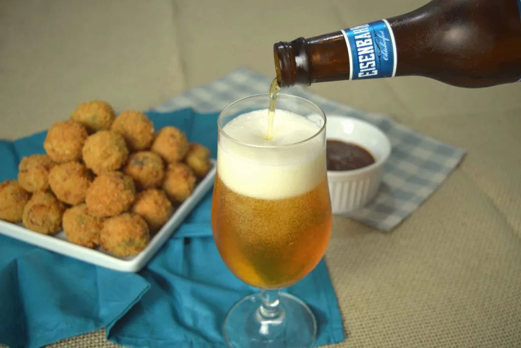 Cerveja Eisenbahn Oktoberfest sendo servida em taça, com bolinhos de aipim empilhados e molho ao fundo