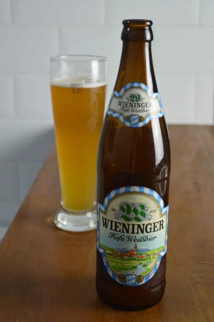 Garrafa de Wieninger Hefe Weissbier ao lado de copo alto com cerveja dourada e colarinho, sobre mesa de madeira