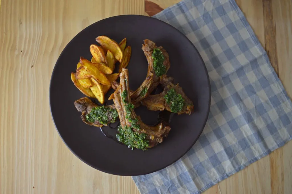 Costelinhas de carré de cordeiro com molho verde de ervas e batatas assadas em prato escuro sobre mesa de madeira