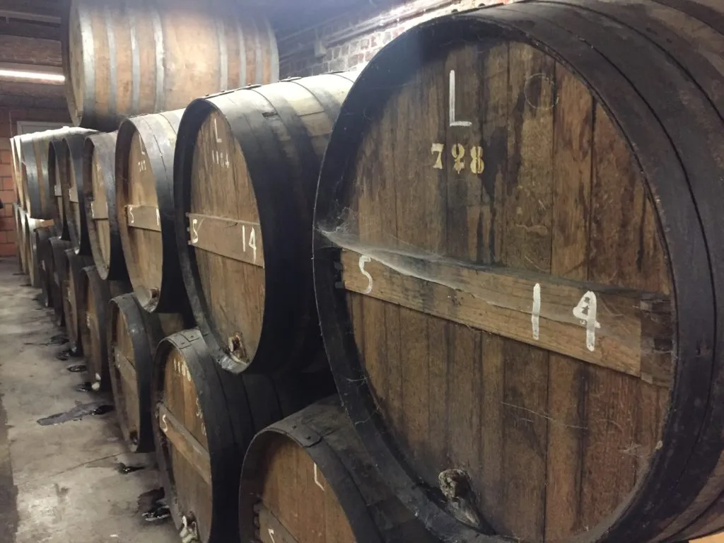 Fileira de grandes barris de madeira empilhados na cervejaria, com números pintados nos tampos