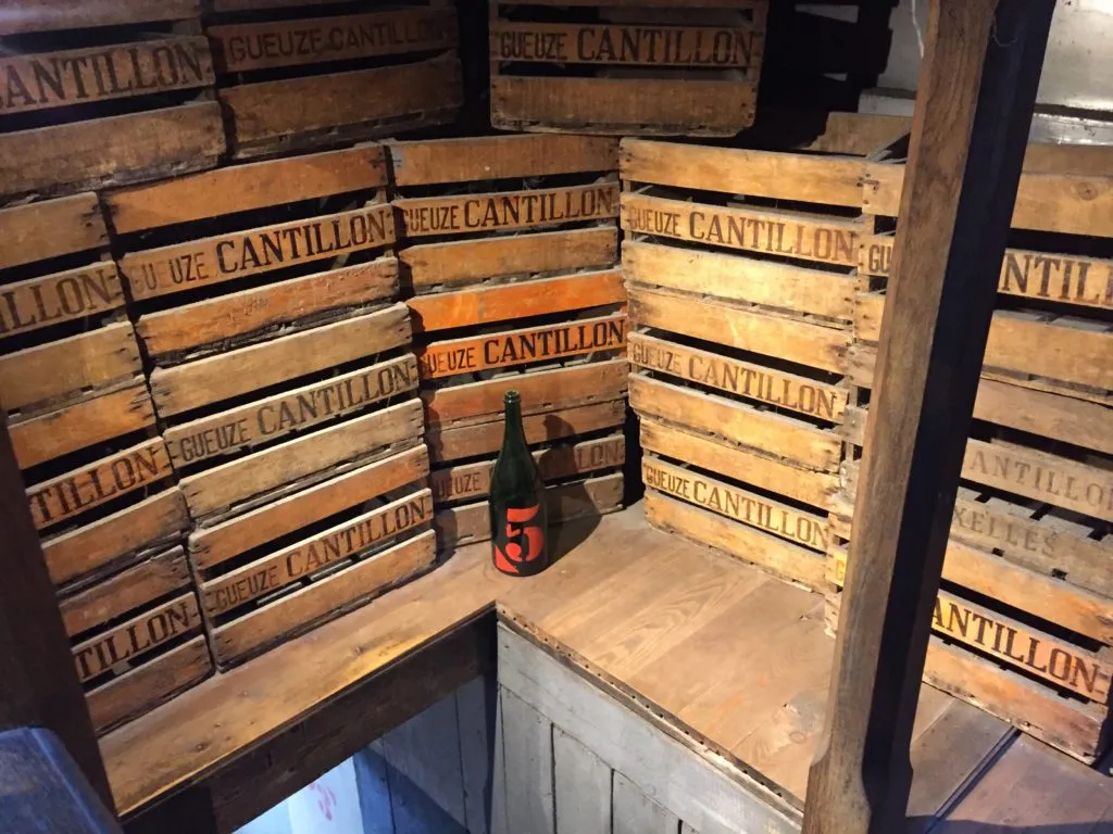Caixas de madeira antigas com a marca Gueuze Cantillon empilhadas, e uma garrafa verde com o número 5