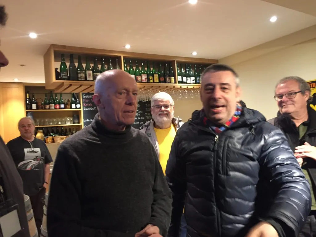 Dois homens conversam em um bar da Cantillon, com prateleiras de garrafas de lambic ao fundo