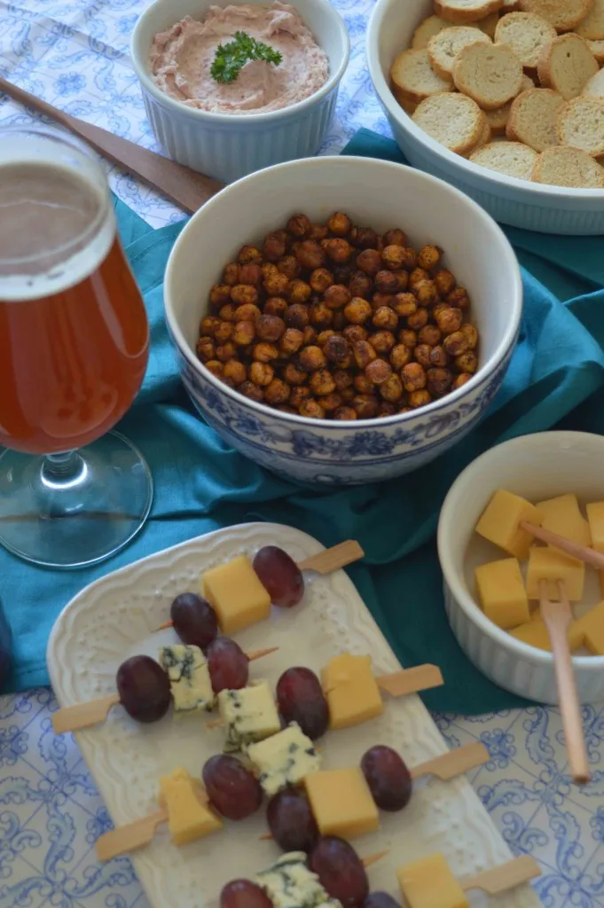 Taça de cerveja âmbar ao lado de tigelas com grão-de-bico assado, torradas, patê e espetinhos de queijo com uvas