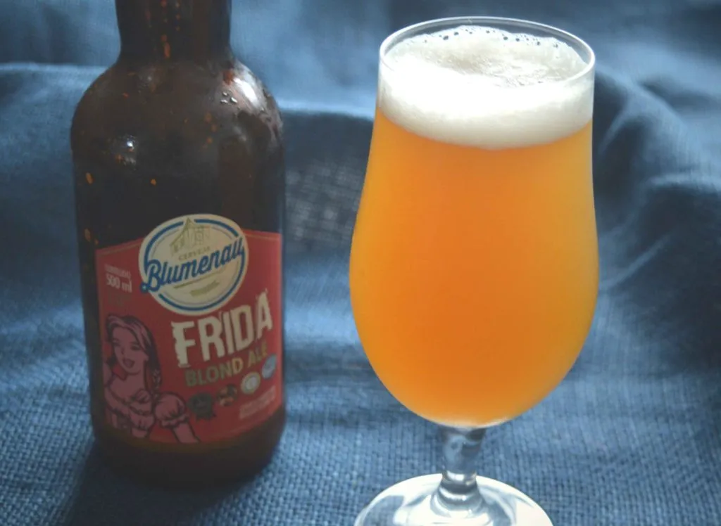 Garrafa de Cerveja Blumenau Frida Blond Ale ao lado de taça com cerveja dourada e espuma