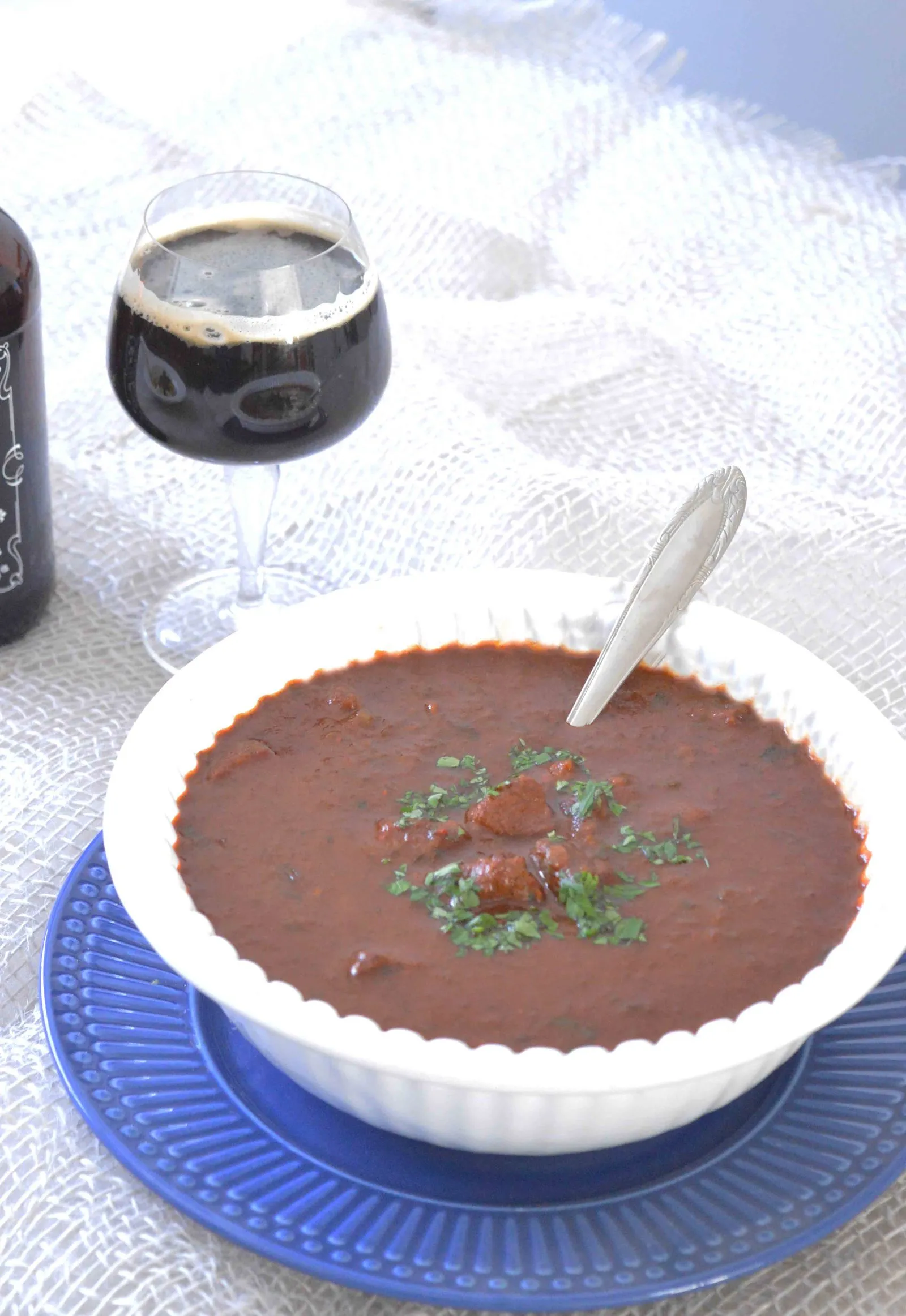 Goulash em tigela branca com salsinha picada, ao lado de taça com cerveja stout escura e colarinho