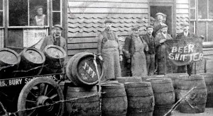 Homens em preto e branco, de época, junto a barris de madeira, com placa Beer Shifter
