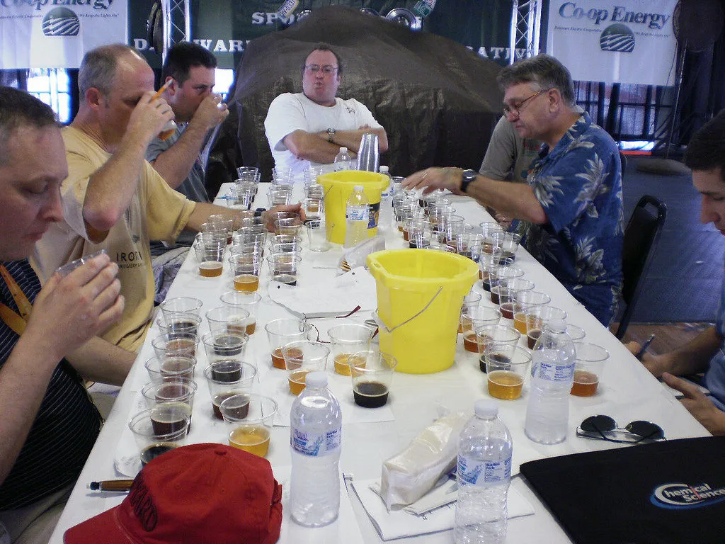 Pessoas provam amostras de cerveja em copos plásticos sobre longa mesa, em evento de avaliação de cervejas