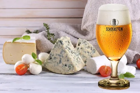 Taça de cerveja loira com colarinho e marca Schornstein, ao lado de queijos azul, brie e mussarela com tomate