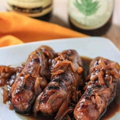 Linguiças grelhadas cobertas com cebolas caramelizadas e molho escuro, num prato, com garrafas de cerveja ao fundo