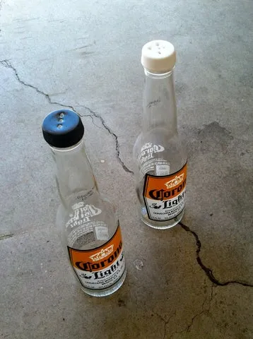 Duas garrafas de cerveja Corona Light vazias com tampas furadas, usadas como saleiro e pimenteiro