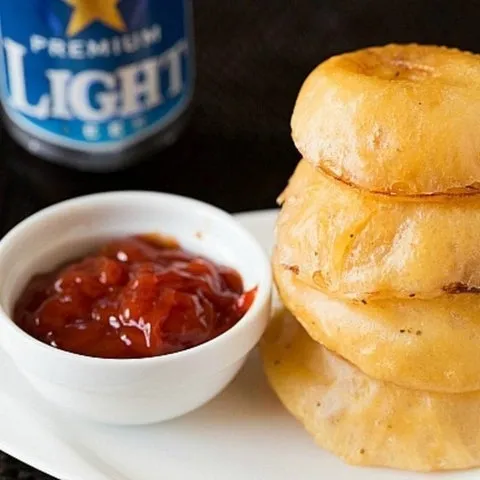 Pilha de onion rings empanados e potinho de molho vermelho, com garrafa de cerveja Light ao fundo