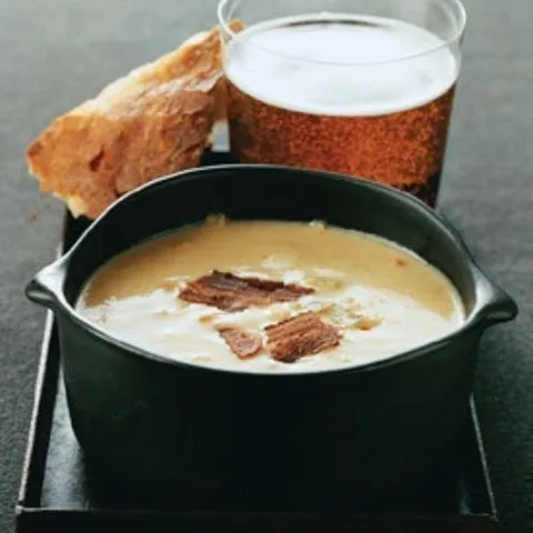Sopa cremosa com bacon em tigela preta, com pão e copo de cerveja ao fundo
