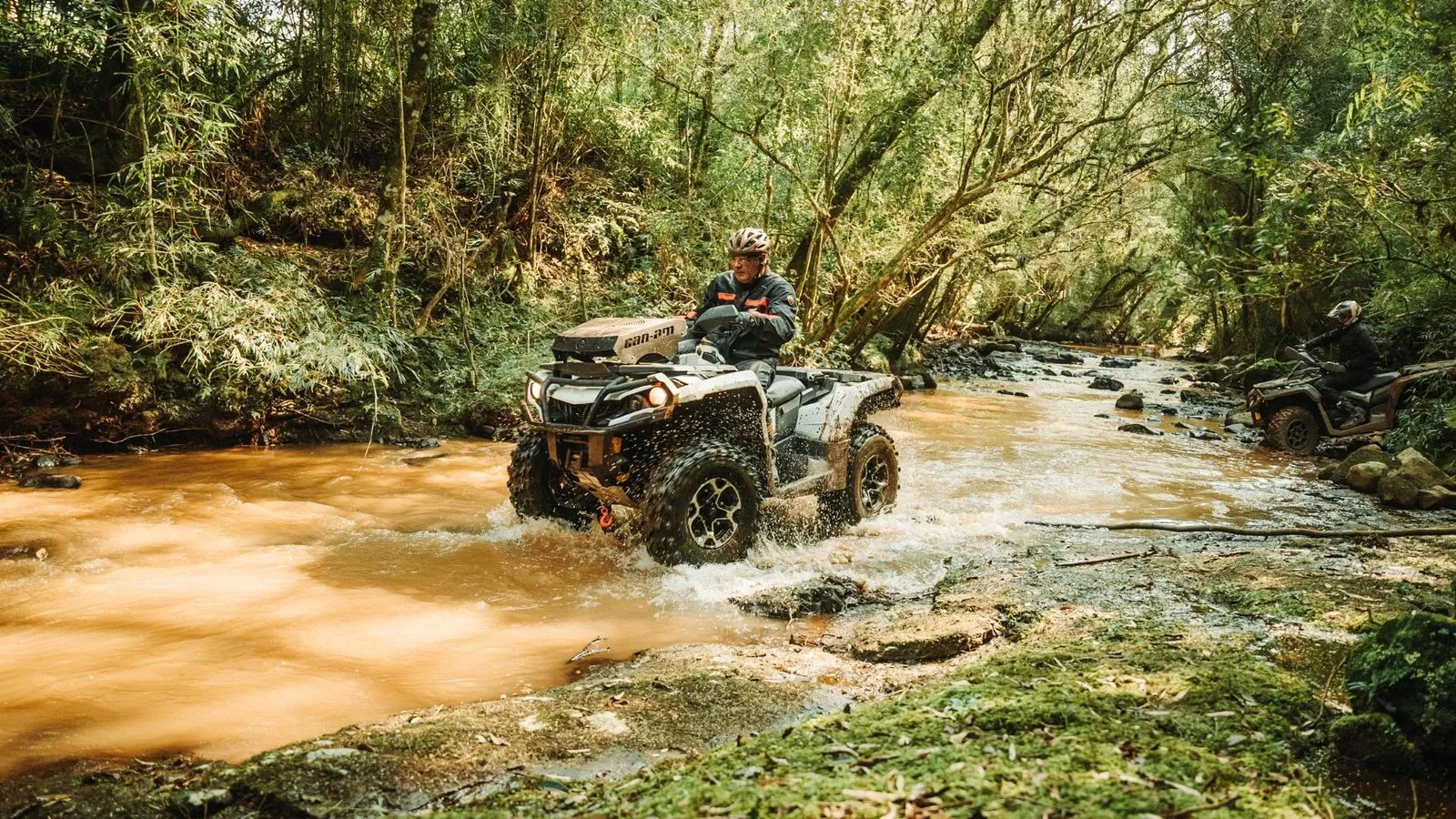 Quadriciclo Can-Am atravessando riacho de águas barrentas em meio à mata, com outro piloto ao fundo