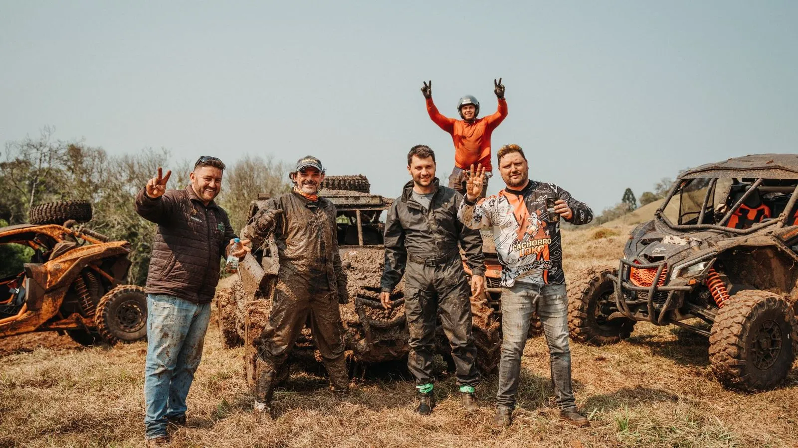 Quatro homens enlameados posam sorrindo em frente a UTVs sujos de barro, com um quinto sobre o veículo