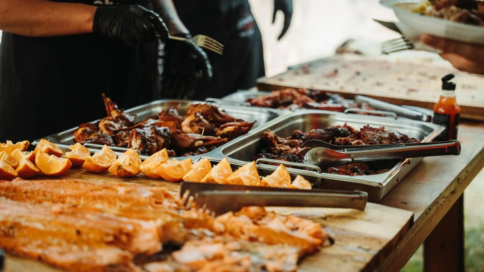 Bufê com travessas de carnes assadas, gomos de laranja e salmão sobre tábua, atendido por pessoas de luvas pretas