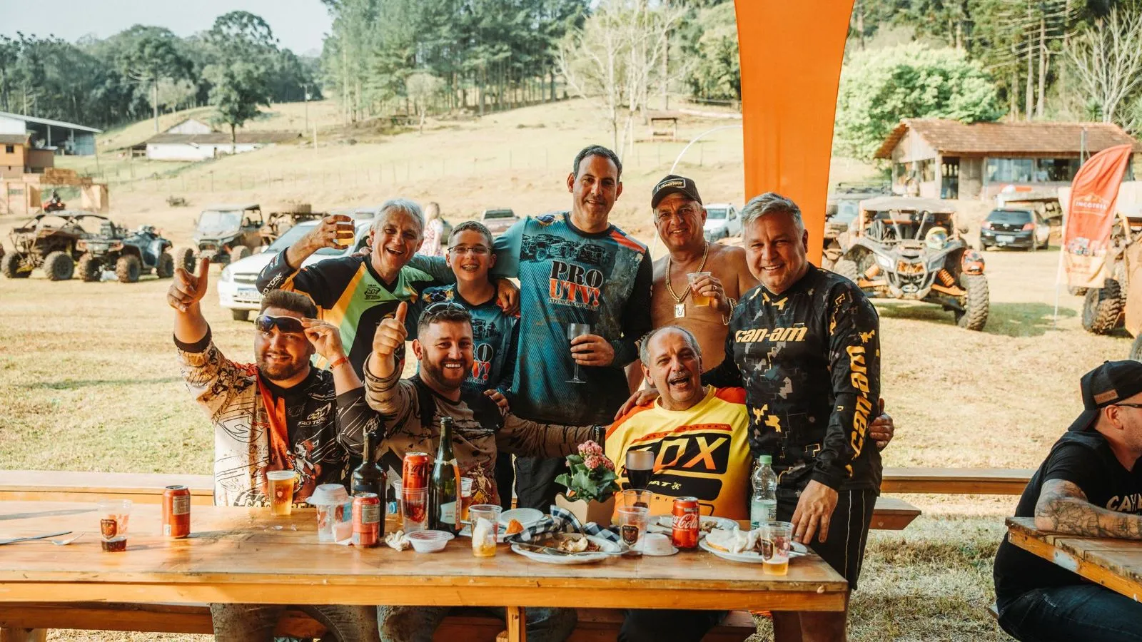 Grupo de participantes do Quadrifest posa à mesa com copos de cerveja, com veículos UTV ao fundo