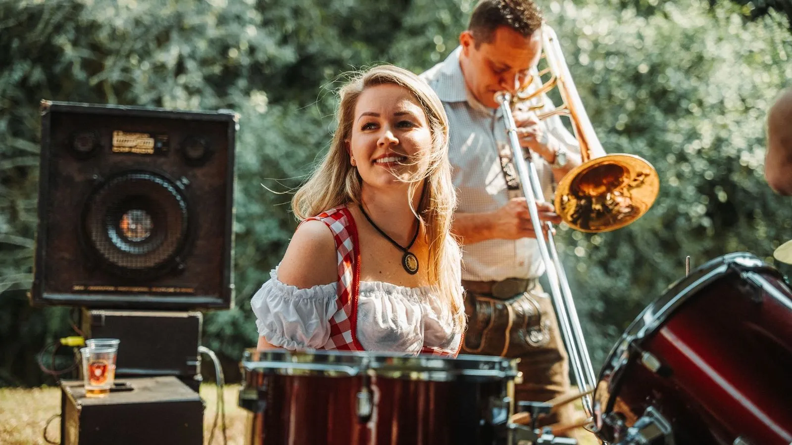 Mulher de traje típico alemão sorri em frente a bateria, com músico tocando trombone ao fundo