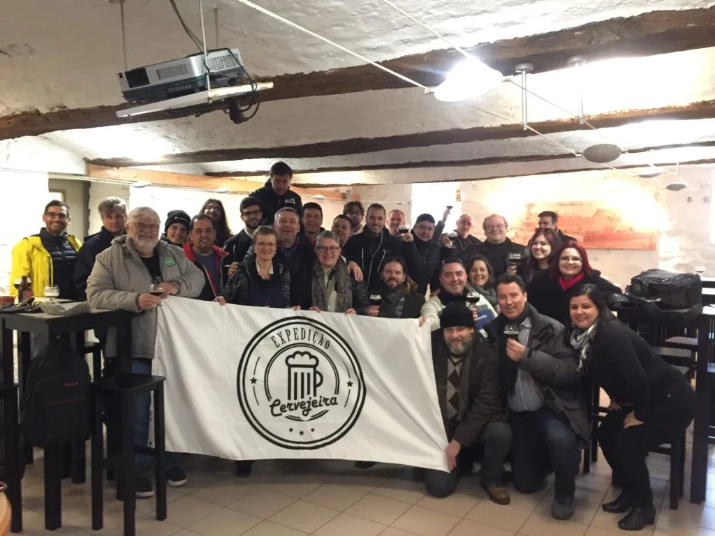 Grupo da Expedição Cervejeira com a bandeira e taças de cerveja em sala com teto de vigas, na Abadia Val-Dieu