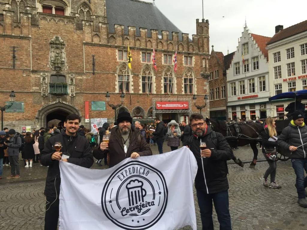 Três homens com copos de cerveja e a bandeira da Expedição Cervejeira na praça de Bruges, em frente ao festival