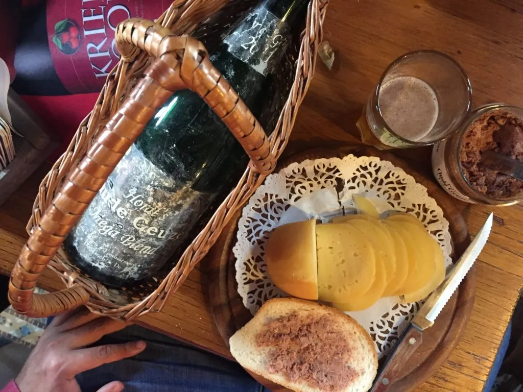 Garrafa de Gueuze em cesto de vime, com tábua de queijo, patê e pão, em mesa de degustação belga