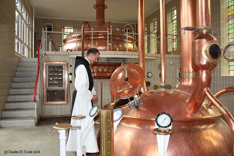 Monge de hábito branco e preto diante de grandes tachos de cobre na sala de brassagem de uma cervejaria trapista