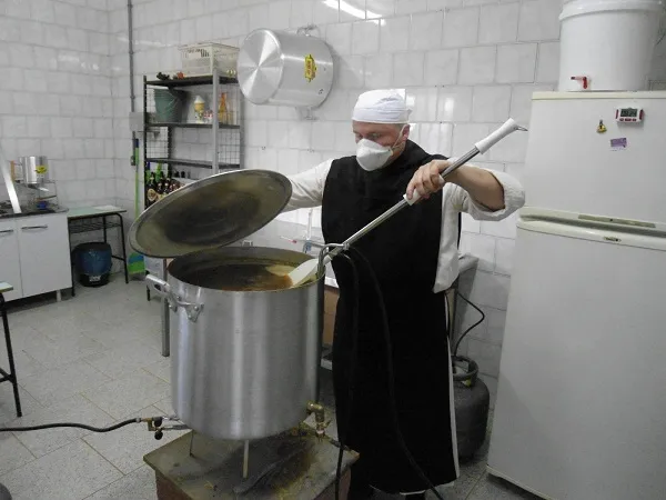 Cervejeiro de avental e máscara mexe o mosto com uma pá em panela grande, em cozinha cervejeira com azulejos