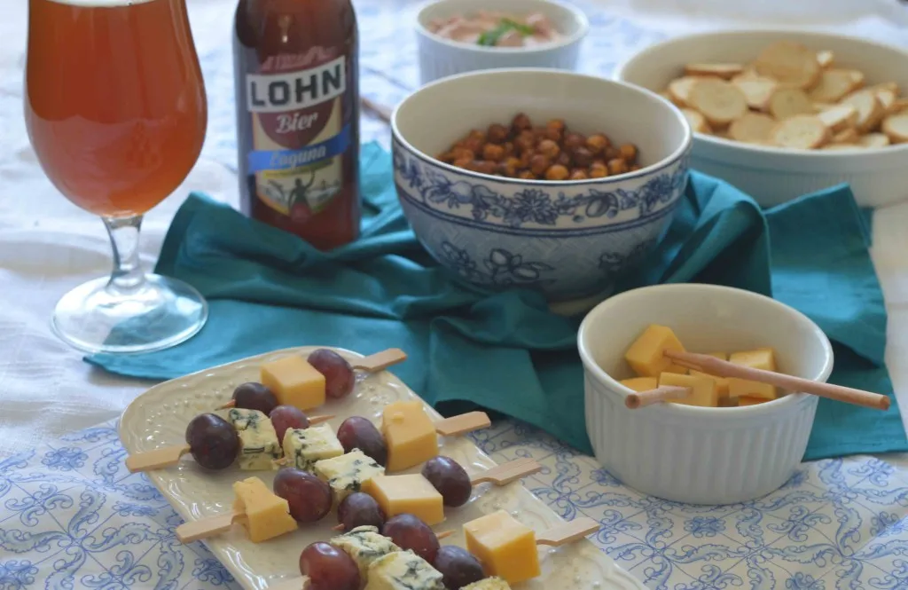 Mesa de petiscos com cerveja Lohn Bier Laguna, grão-de-bico crocante, torradinhas e espetinhos de queijo e uva