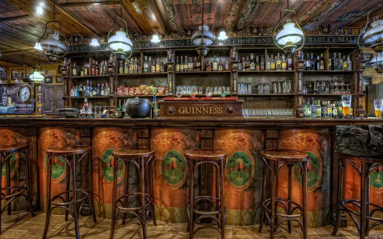 Balcão de pub inglês em madeira escura, com banquetas, prateleiras de garrafas, lustres e torneira Guinness