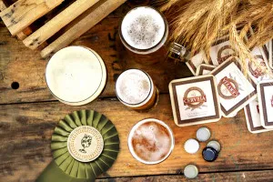 Mesa de madeira com copos de cerveja, porta-copos, rosácea de premiação e espigas de cevada