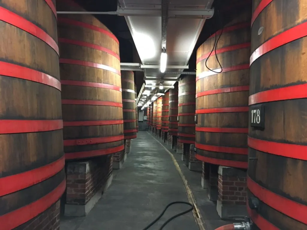Corredor entre grandes dornas de madeira com aros vermelhos na cervejaria Rodenbach